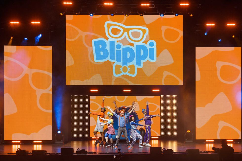 A big stage with "Blippi" displayed on the middle of three projection screens. Blippi, wearing a blue shirt, suspenders, orange glasses and hat, entertains audience members alongside his fellow cast.