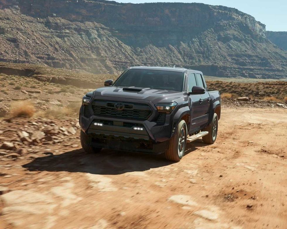 A black manual Toyota Tacoma driving across a red, rocky landscape.
