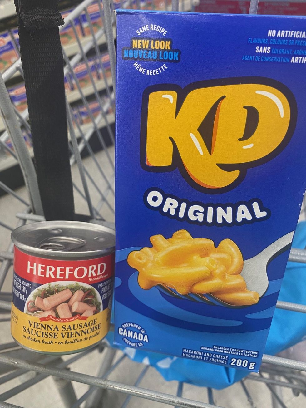 A blue box of KD mac and cheese and a can of Vienna sausage in a shopping cart.