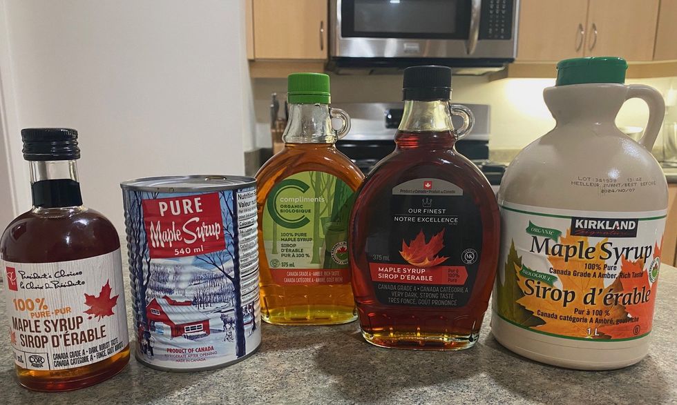 A bottle of President's Choice maple syrup, a can of Decacer maple syrup, a bottle of Compliment's maple syrup, a bottle of Our Finest maple syrup and a bottle of Kirkland Signature maple syrup.
