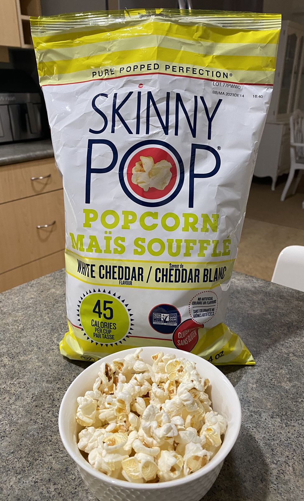 A bowl filled with white cheddar flavour popcorn with a bag of popcorn behind it on a kitchen counter.