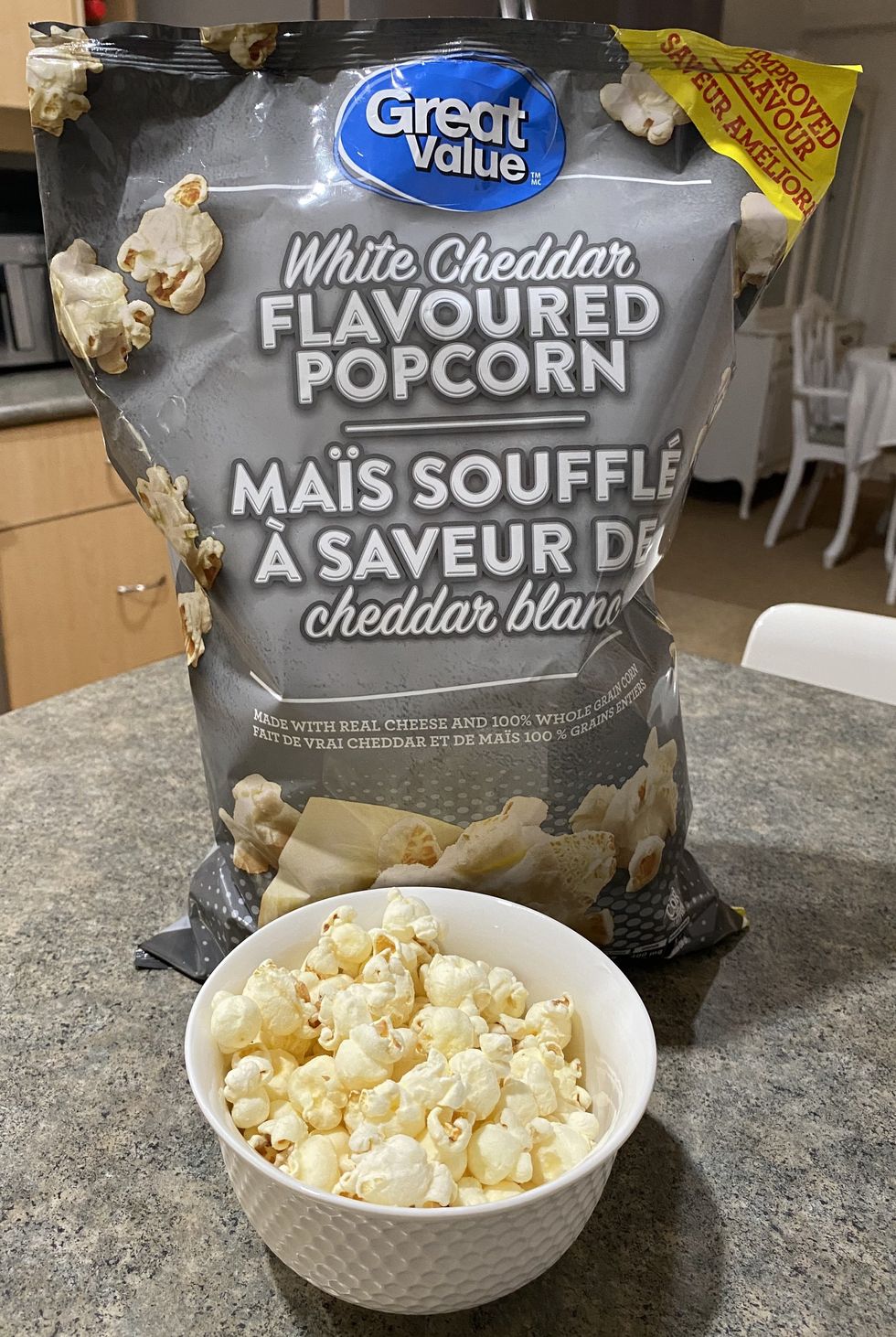A bowl filled with white cheddar flavour popcorn with a bag of popcorn behind it on a kitchen counter.
