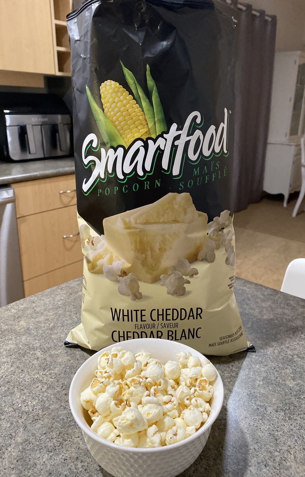 A bowl filled with white cheddar flavour popcorn with a bag of popcorn behind it on a kitchen counter.