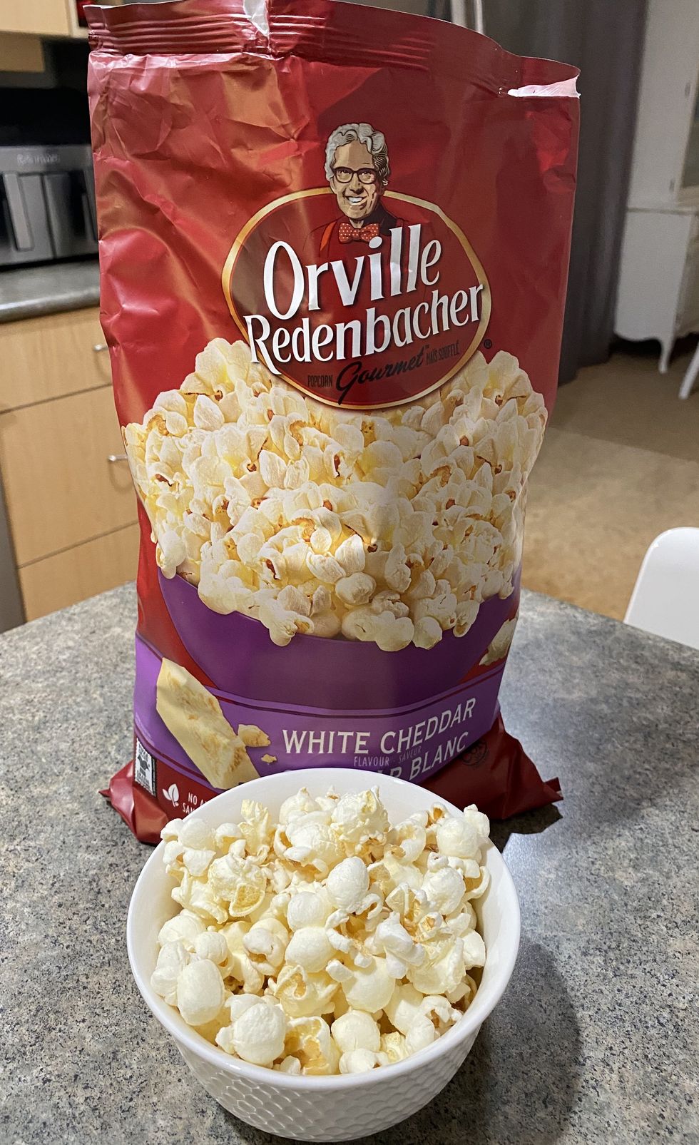 A bowl filled with white cheddar flavour popcorn with a bag of popcorn behind it on a kitchen counter.