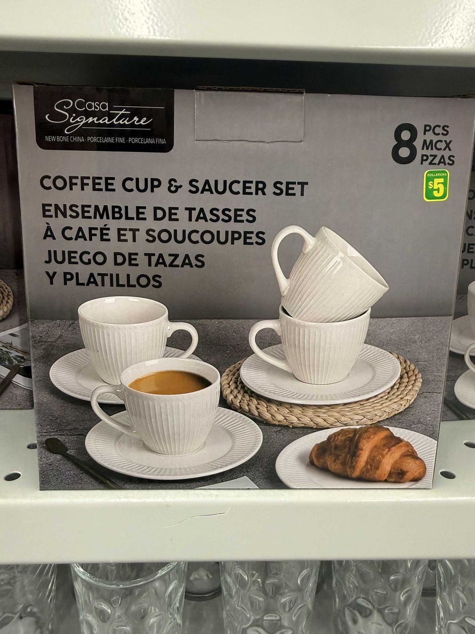 A box with white coffee cups and matching plates with a croissant on one of them on a store shelf.