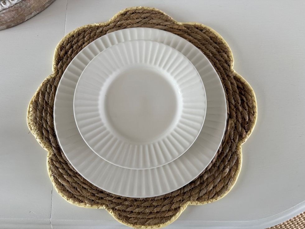 A braided straw flower-shaped placemat on a white table with two white dishes stacked on top of it.