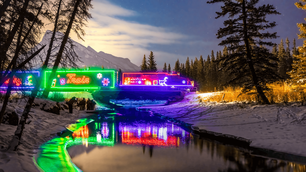 A brightly lit holiday train rolls through a snowy mountain landscape at dusk.