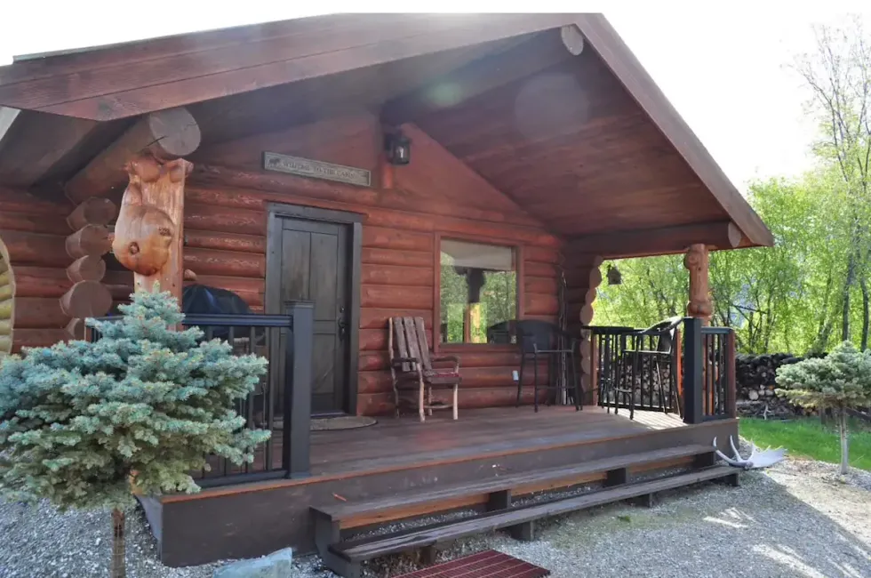 A brown log cabin with a black door and chairs on the porch and trees around it.