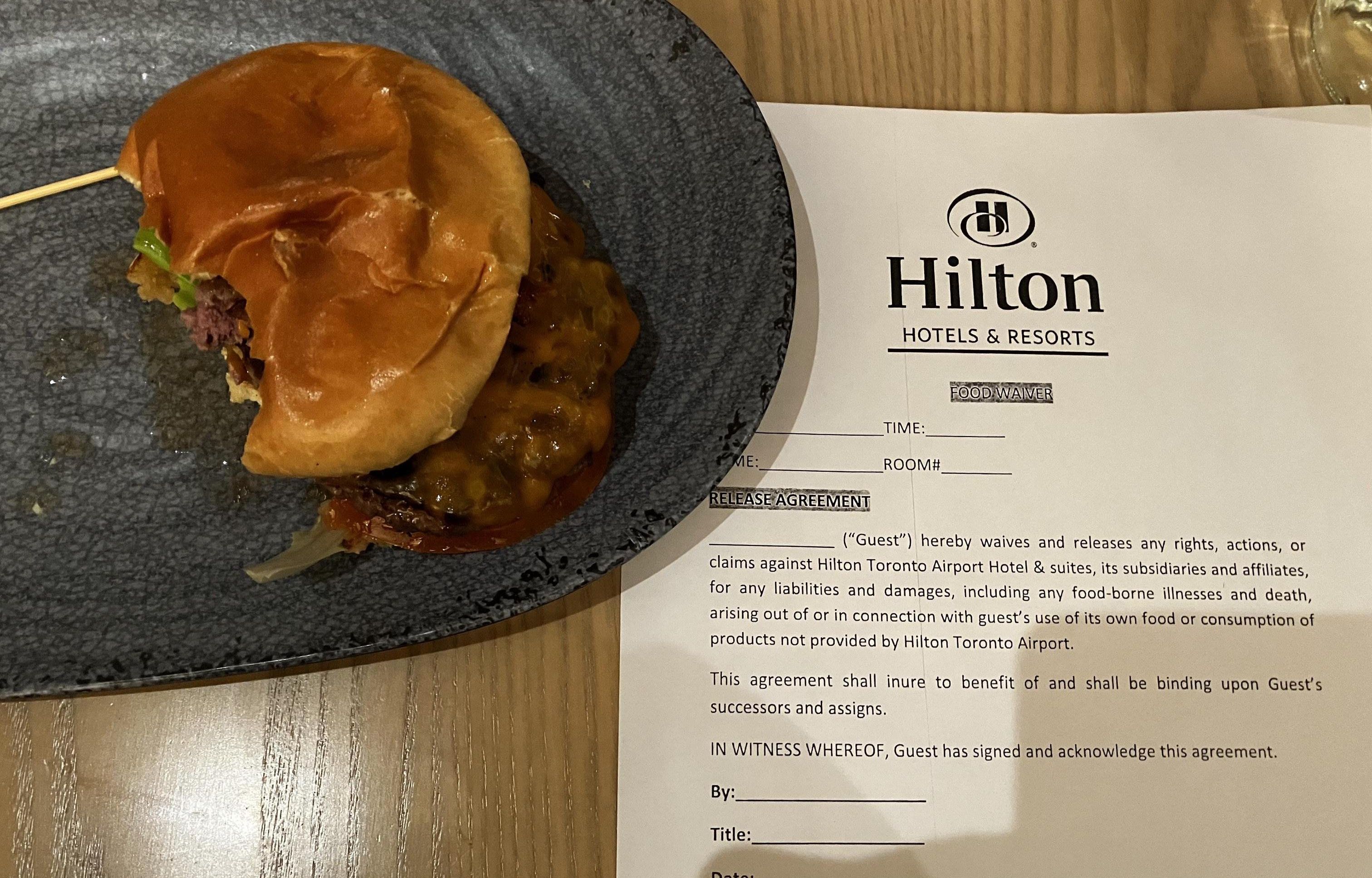 A burger on a plate with a bite taken out of it and a food waiver form from the Hilton hotel.