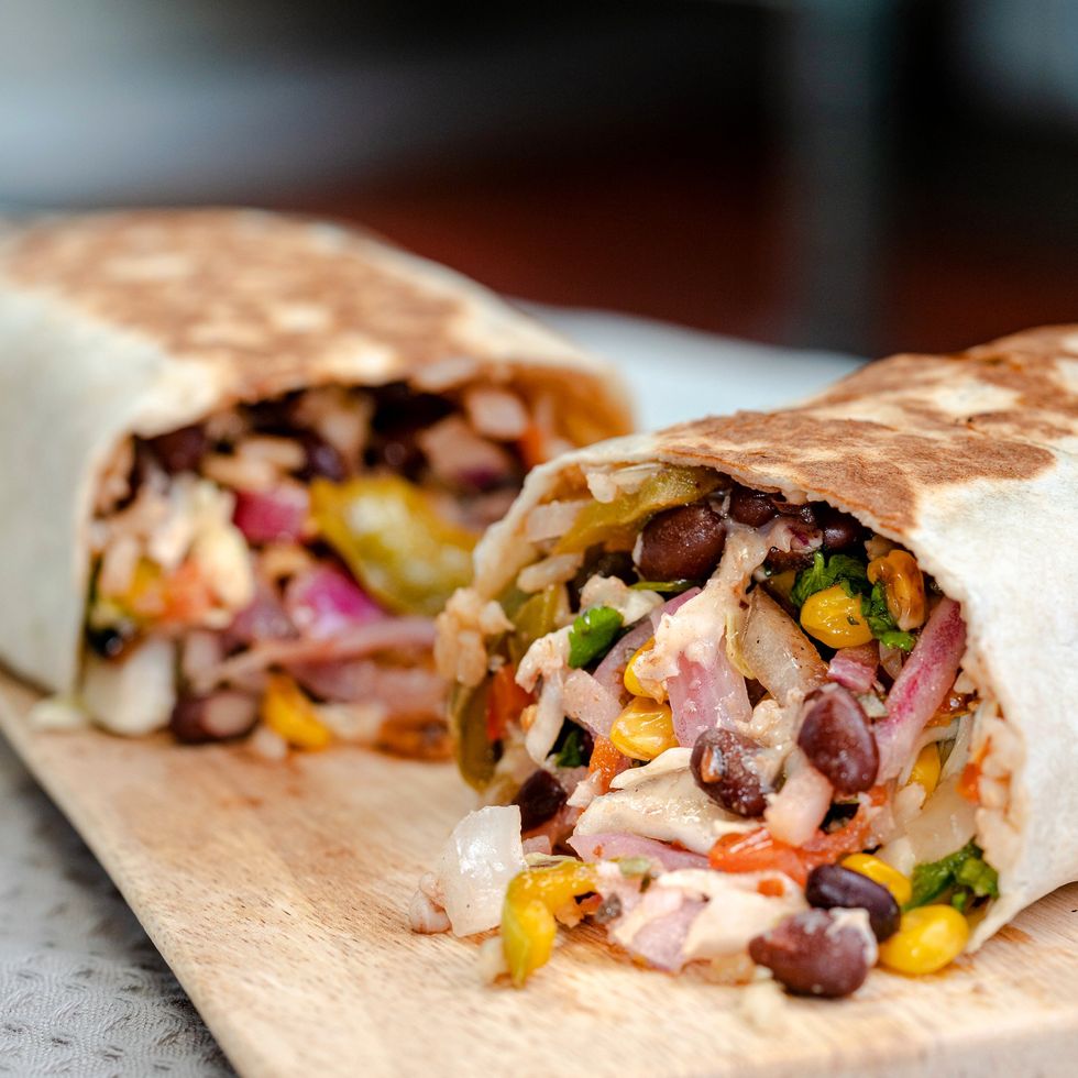 A burrito cut in half with beans,  revealing beans, corn, salsa and cheese inside, resting on a wooden board. 