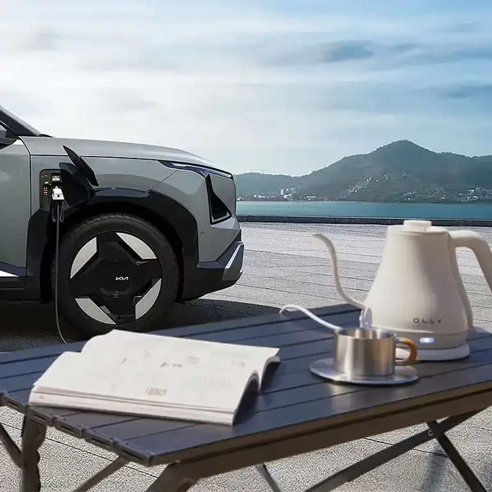 A cable connects the Kia EV5 to an electric kettle on a camp table next to a cup and open book.