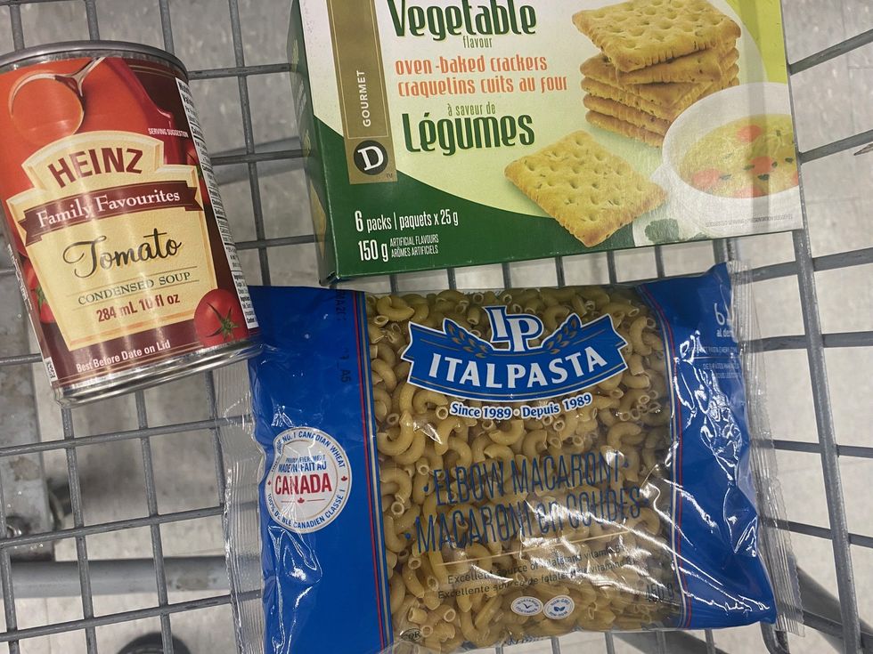 A can of Heinz Tomato condensed soup, a box of vegetable flavour crackers and a bag of elbow macaroni in a shopping cart.