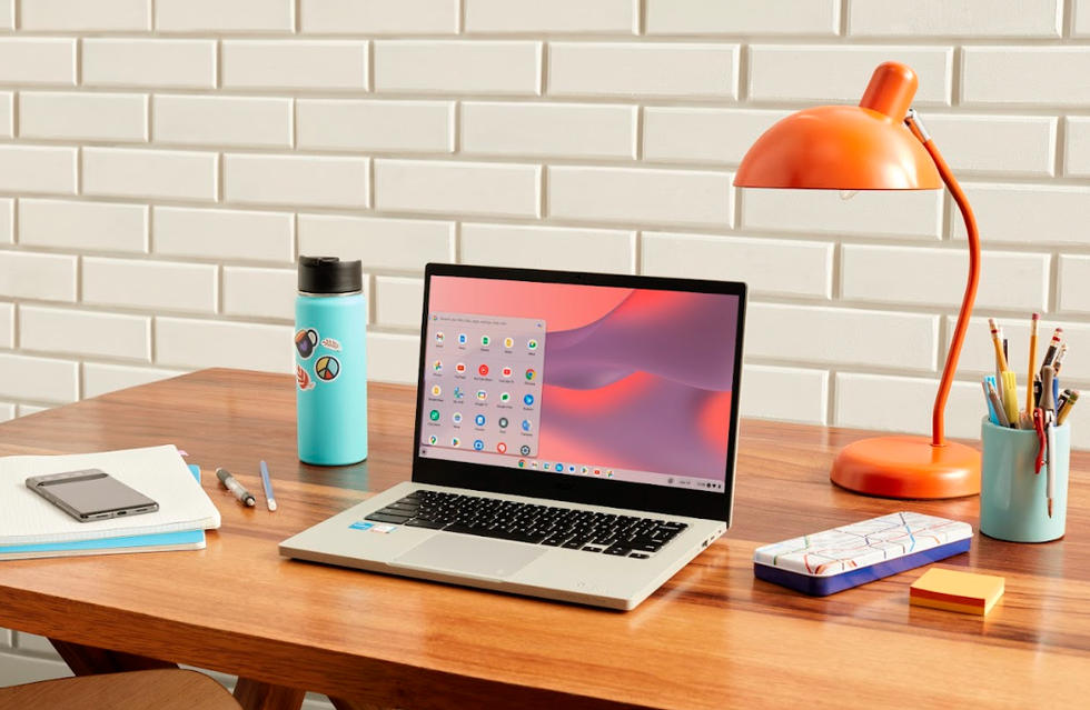 A Chromebook laptop sits open on a desk with a blue water bottle, notebook and phone to the left and an orange lamp, sticky notes and pen holder on the right.