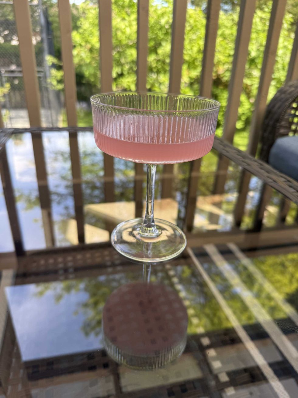 A clear glass with pink liquid inside sitting on a glass table outside.