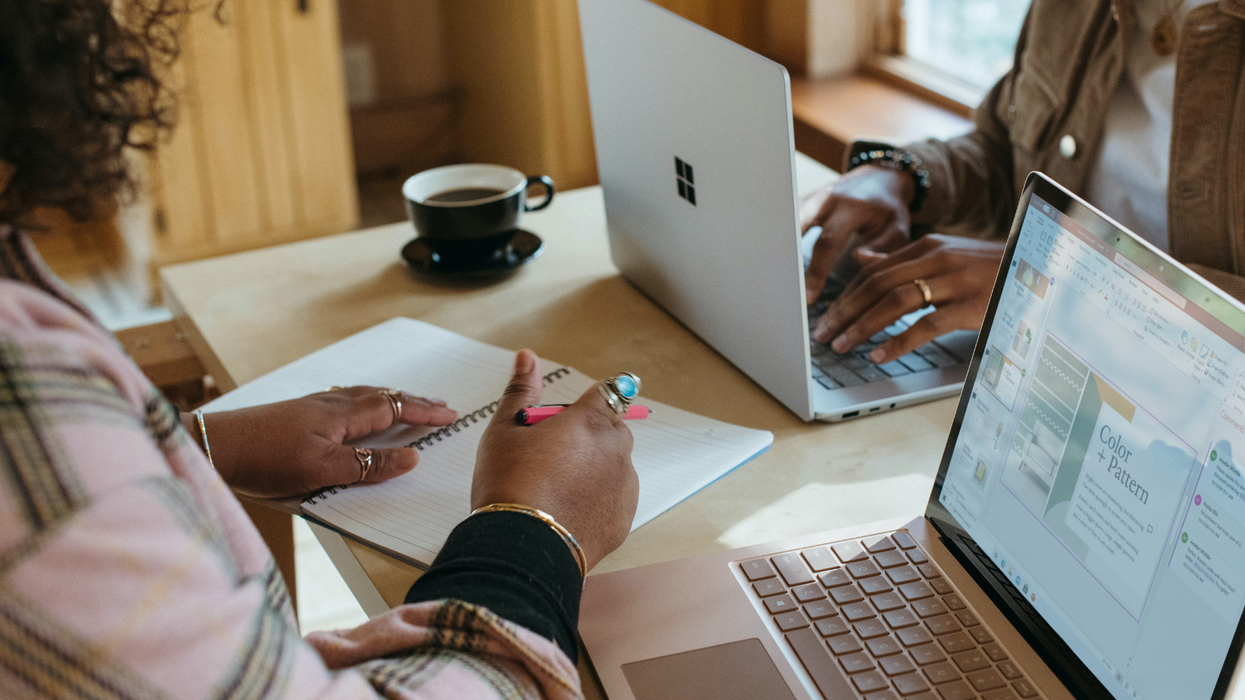 A close shot of a desk shared by two people working on laptops, one person is holding a pen about to write in a notebook.