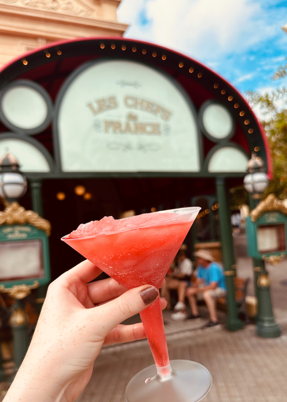 A close-up of a frozen red cocktail held in front of the "Les Chefs de France" restaurant at EPCOT in Disney World.
