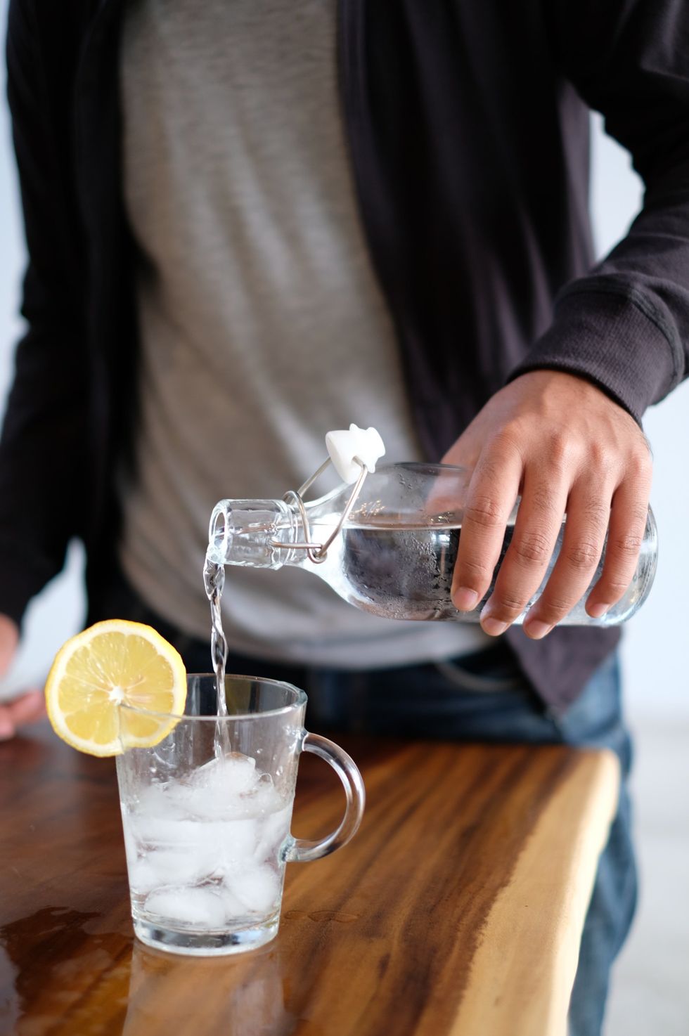 A close up of an ice-filled glass mug with a lemon wedge on the rim. A person is filling the cup with water from a clear glass bottle.