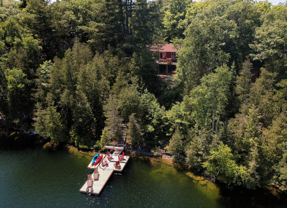 A cottage is seen on a hill by an emerald lake with a dock leading to the water.