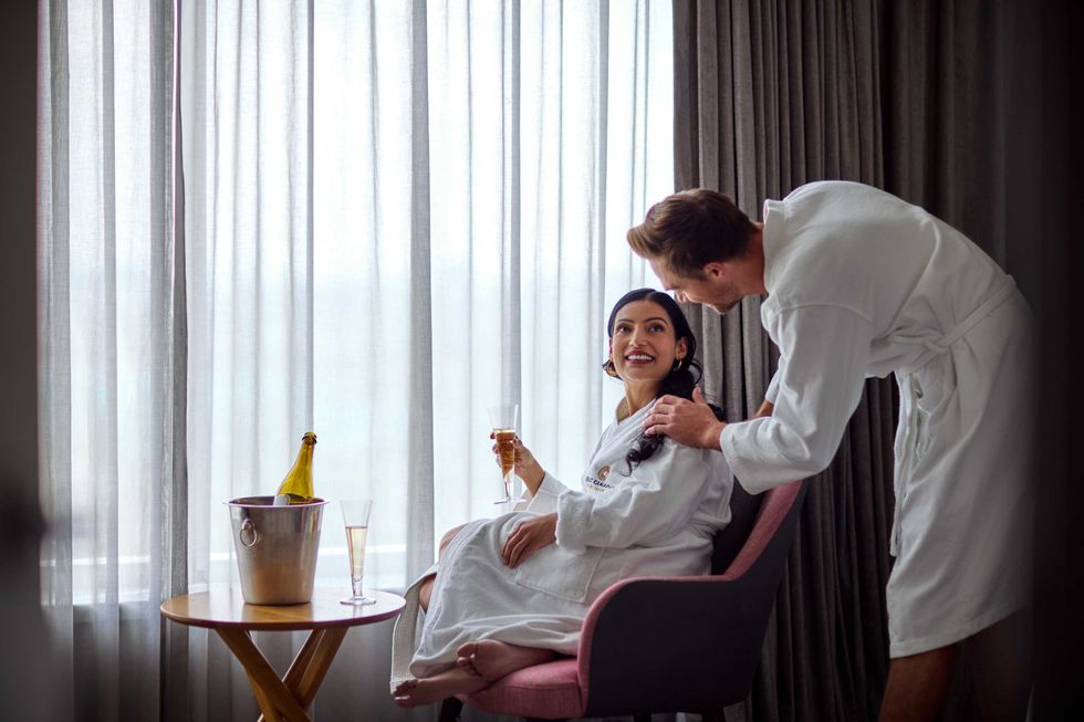 A couple in a hotel room wearing robes sip champagne in front of a sheer-curtained window.