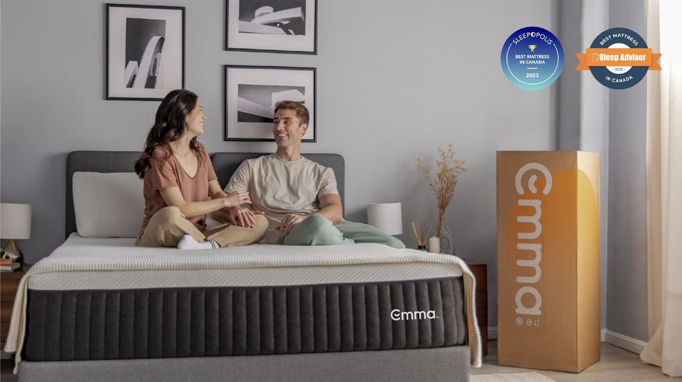 A couple sit on a bed with an Emma Sleep Hybrid Comfort mattress. An Emma Sleep box stands on its end in the corner and the couple are smiling at each other.