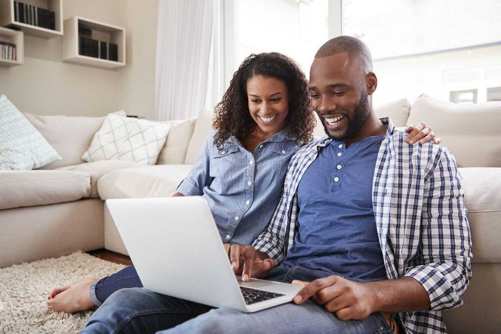 A couple sitting on the floor of their home, smiling at a laptop.