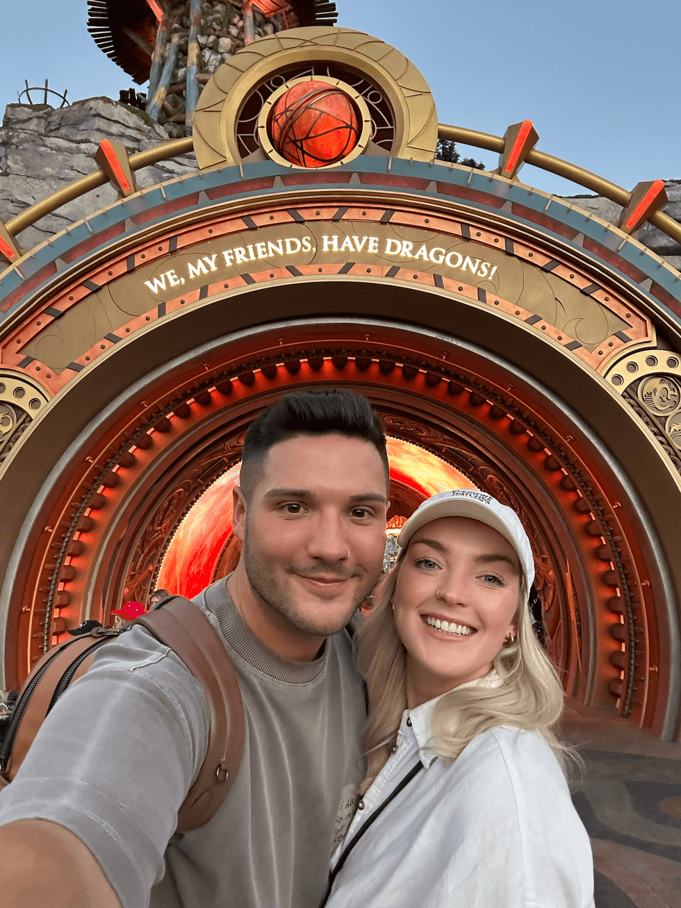 A couple smiling in a selfie in front of the circular portal entrance to How to Train Your Dragon: Isle of Berk at Epic Universe, with the inscription "We, my friends, have dragons!" arched above them in glowing orange light.