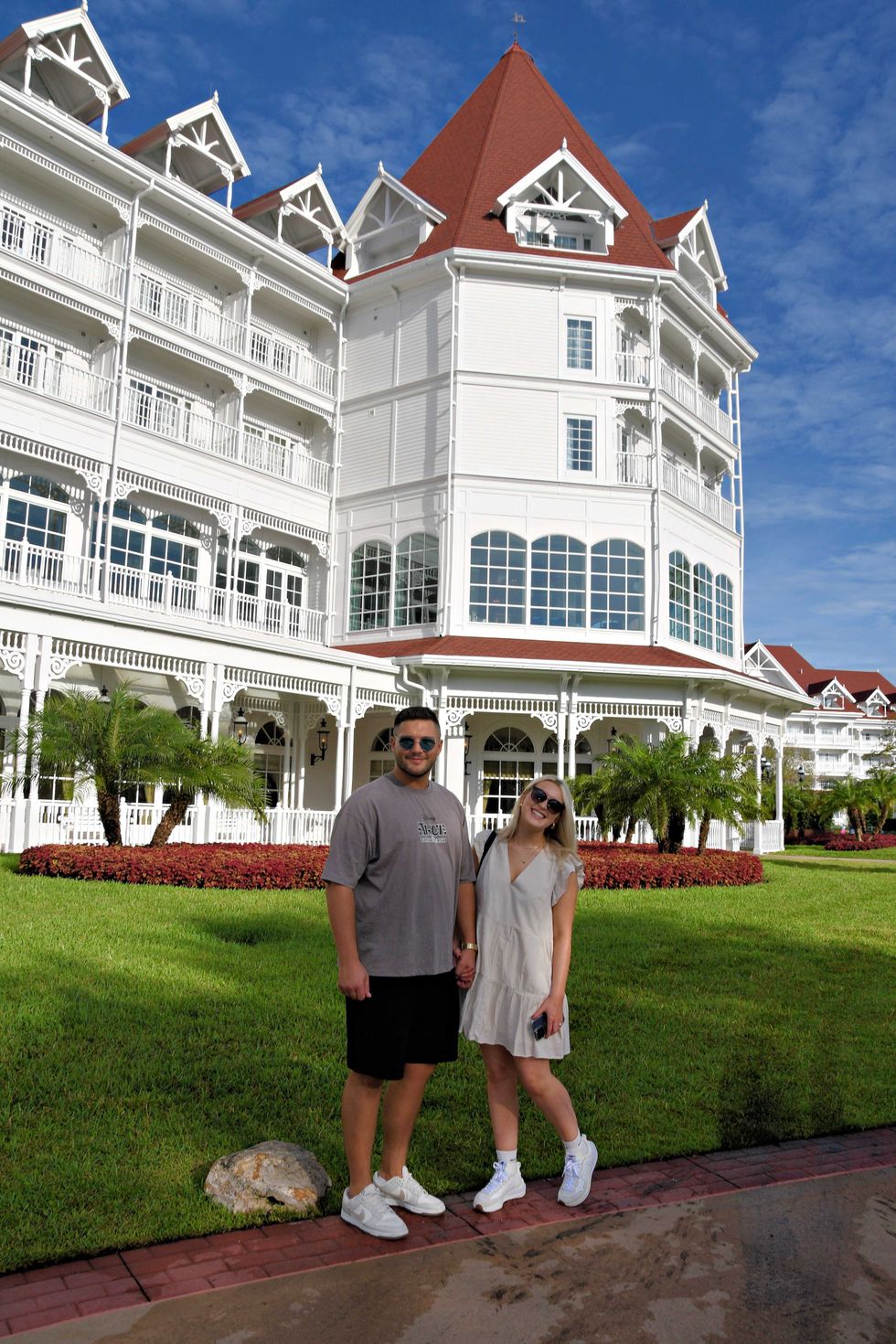 A couple smiling in front of the white Victorian exterior of Disney's Grand Floridian Resort & Spa, with red shingle roofs, ornate gingerbread trim, and manicured gardens behind them.