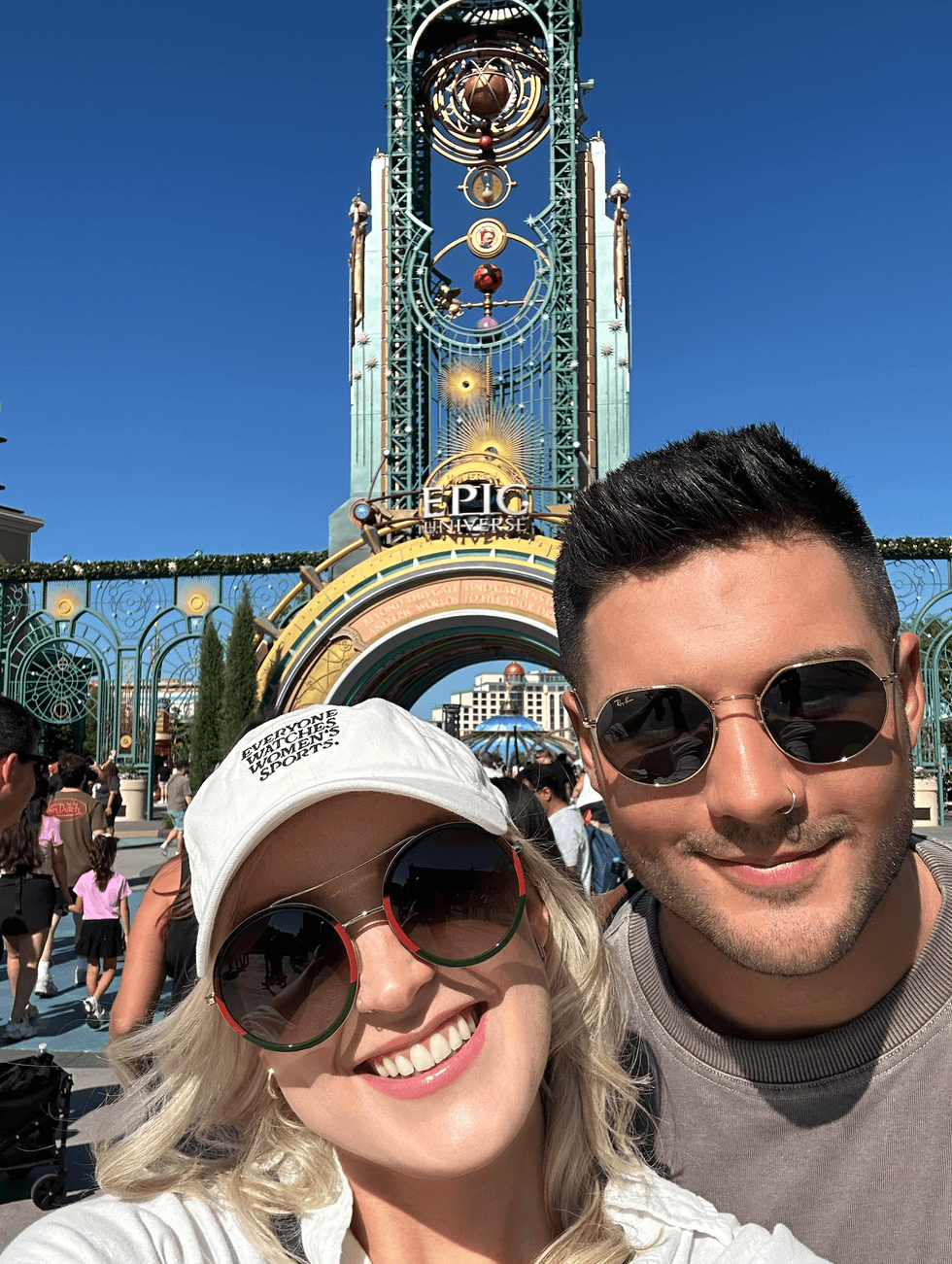 A couple taking a selfie in front of the ornate art deco entrance gates of Universal's Epic Universe, with the towering "Epic Universe" sign and Helios Grand Hotel visible behind them under a bright blue sky.