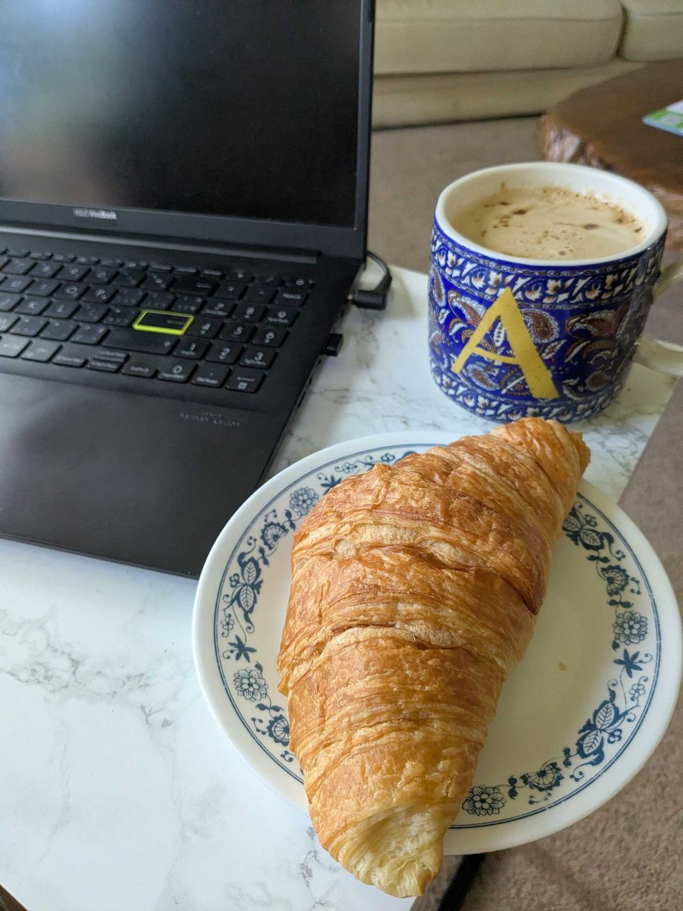A croissant on a plate and a cup of copy beside a laptop computer on a table