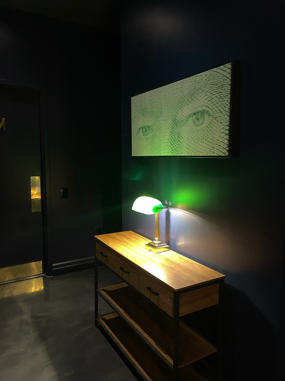 A desk with a banker style lamp and painting hung above it.