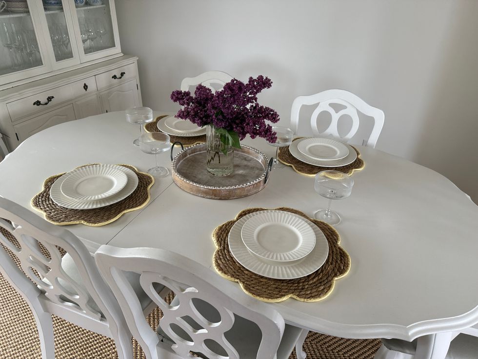 A dining room table with four chairs and a place setting on tip, including four placemats, plates, cups and a centre piece of flowers and a tray.