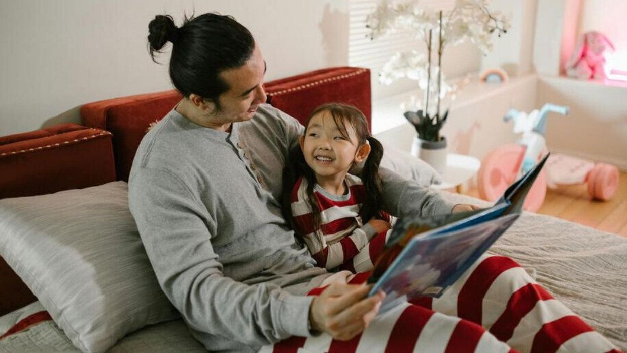 A father and daughter in matching white and red striped pyjamas read a book together on a bed.