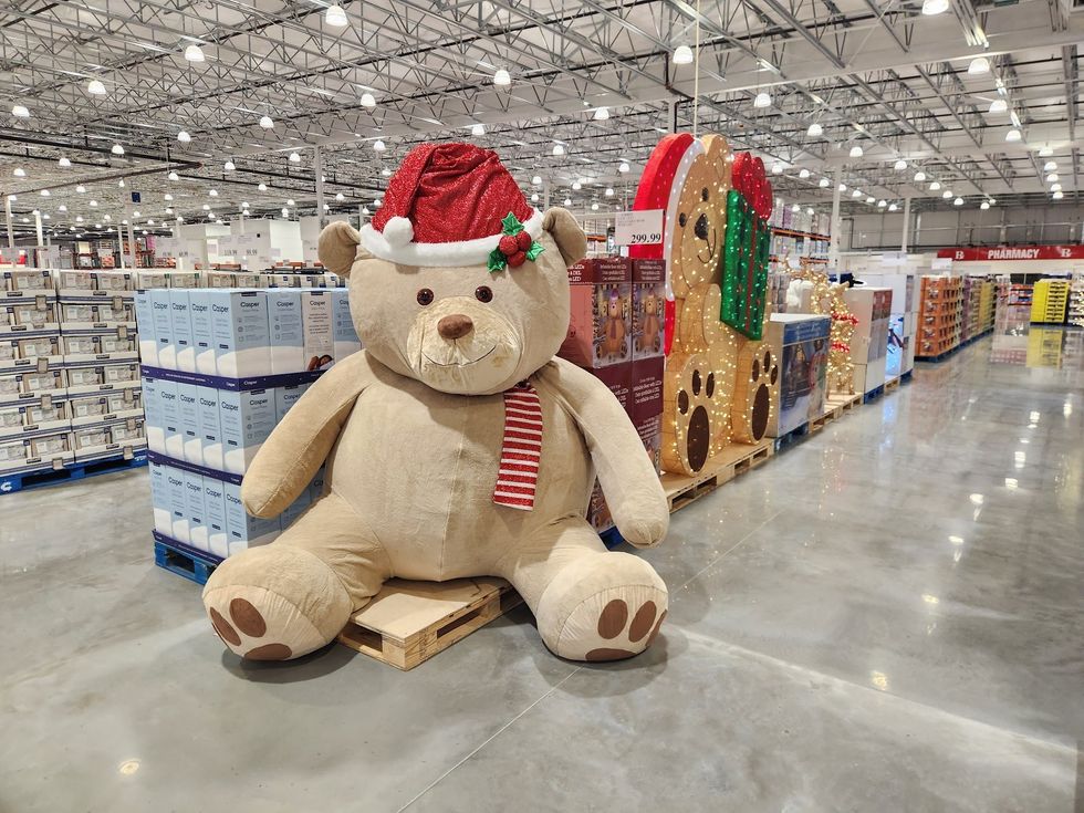 A giant teddy bear with a red Santa hat and festive scarf sits on display beside illuminated holiday decorations in the Costco\u2019s seasonal section.