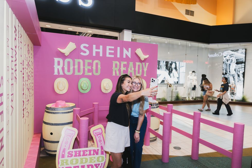 A girl in a black top and white shorts takes a selfie with a friendin a blue shirt in front of a sign that says "SHEIN Rodeo Ready"