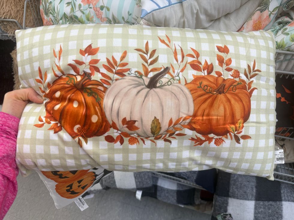 A green and white checkered pillow with two orange and one white pumpkin surrounded by leaves.