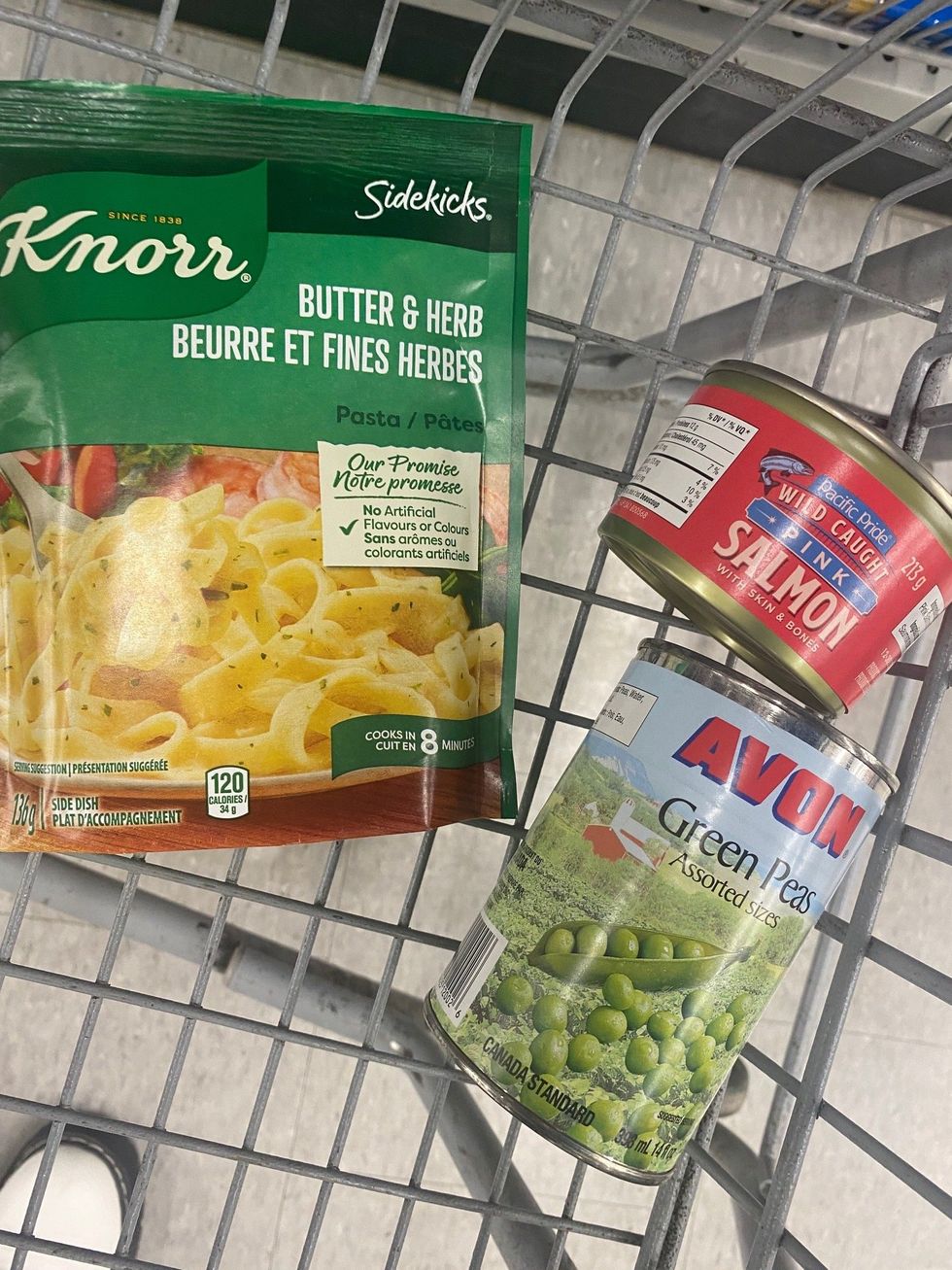 A green bag of Knorr Sidekicks pasta, a red can of salmon and a can of green peas in a shopping cart.