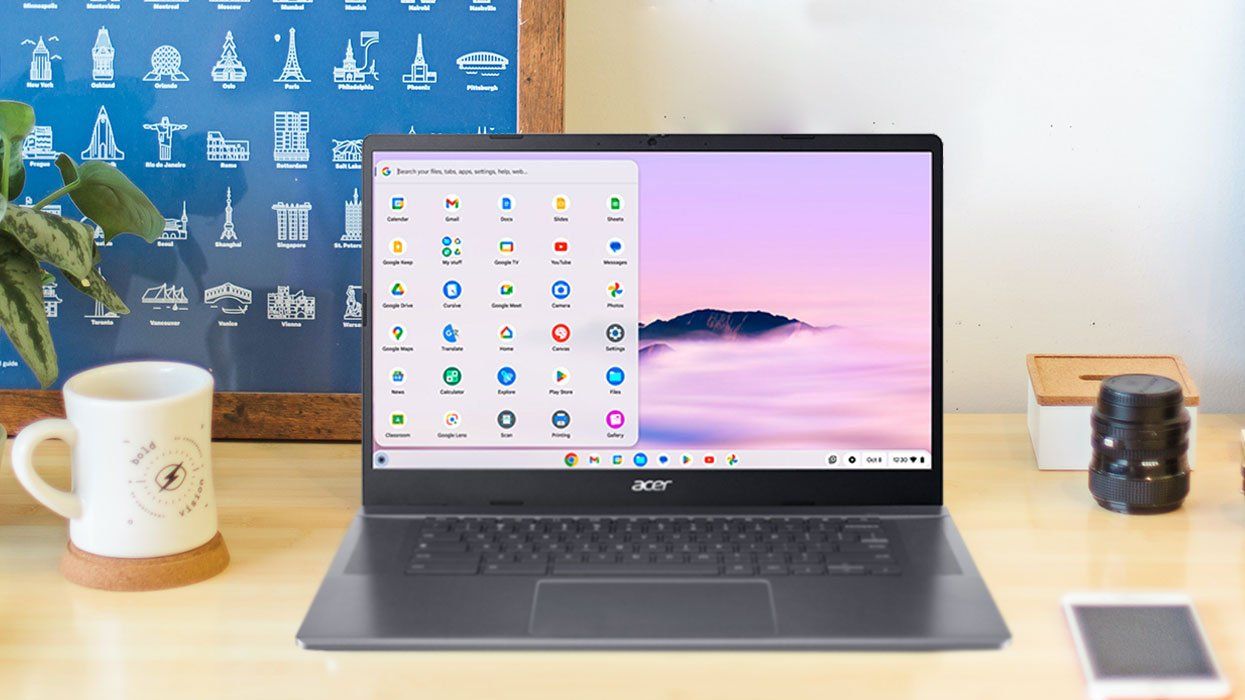 A grey Acer Chromebook Plus 515 laptop sits on a desk facing the camera with a phone and camera lens to the right of it and a coffee mug, plant, and framed pictures to the left.