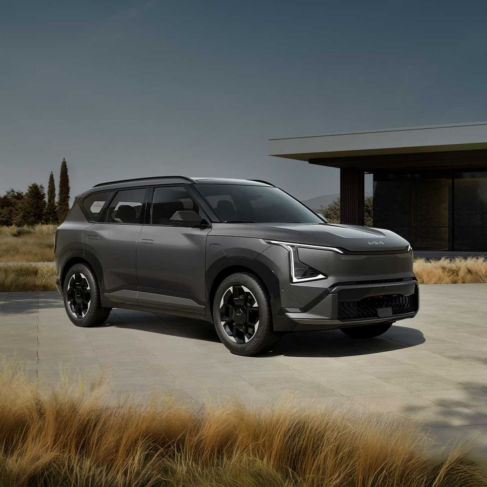 A grey Kia EV5 parked on pavers in front of a modern house and grassy field.
