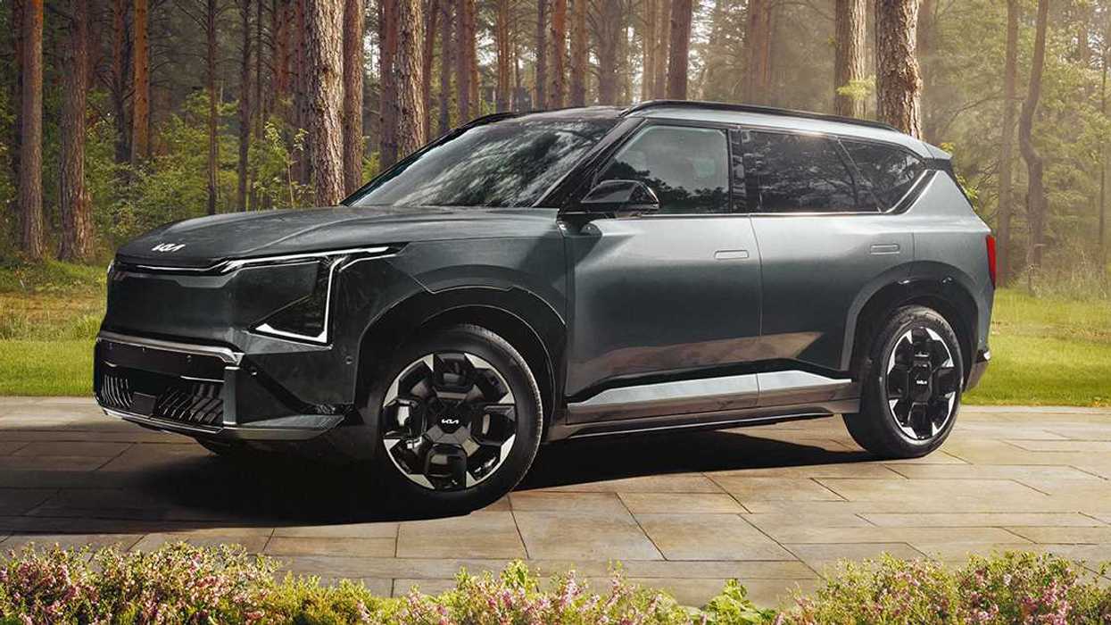 A grey Kia SUV parked on a paved driveway with trees behind.