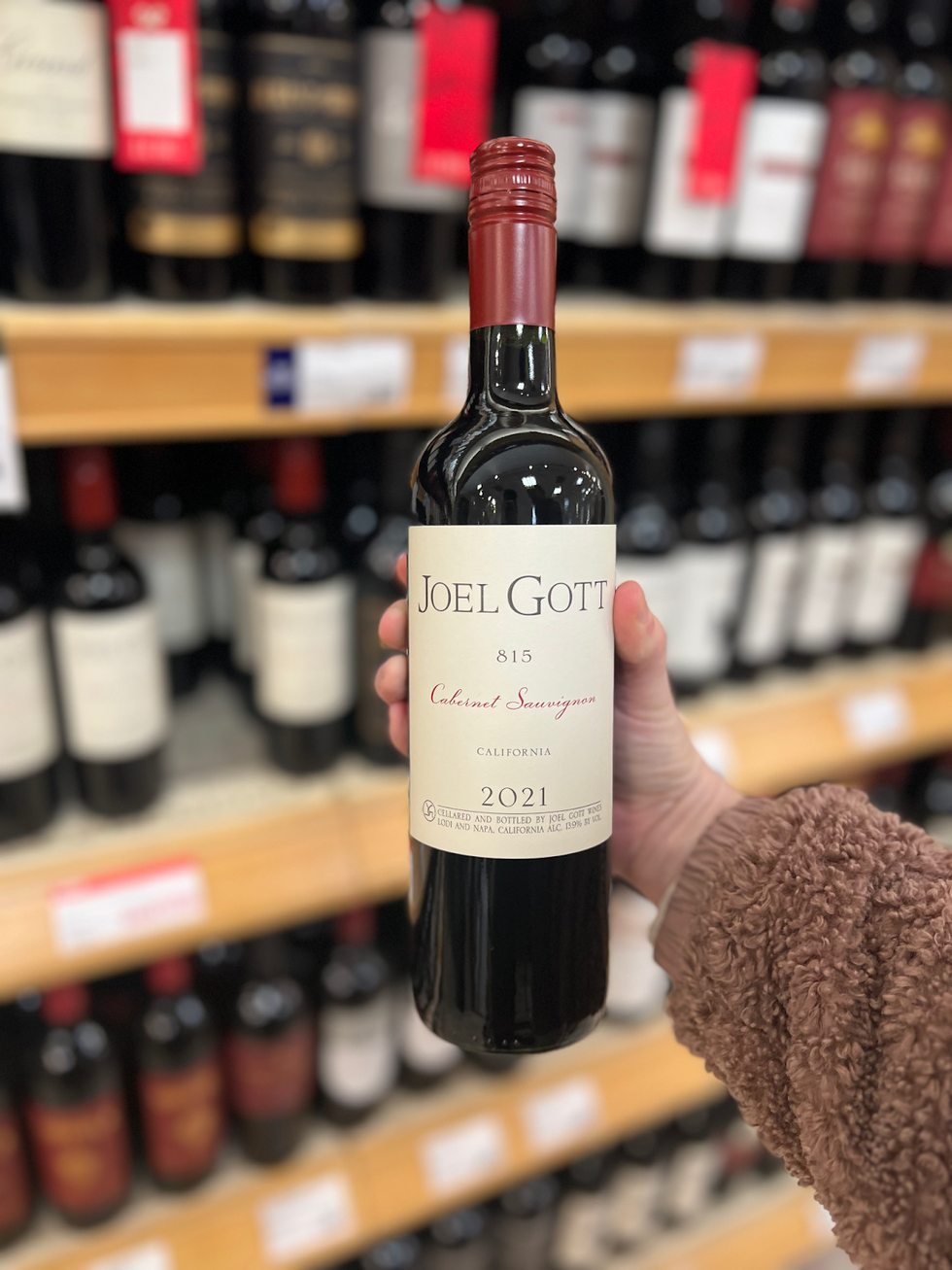 A hand holding a bottle of Joel Gott 815 Cabernet Sauvignon in front of a rack of wine.