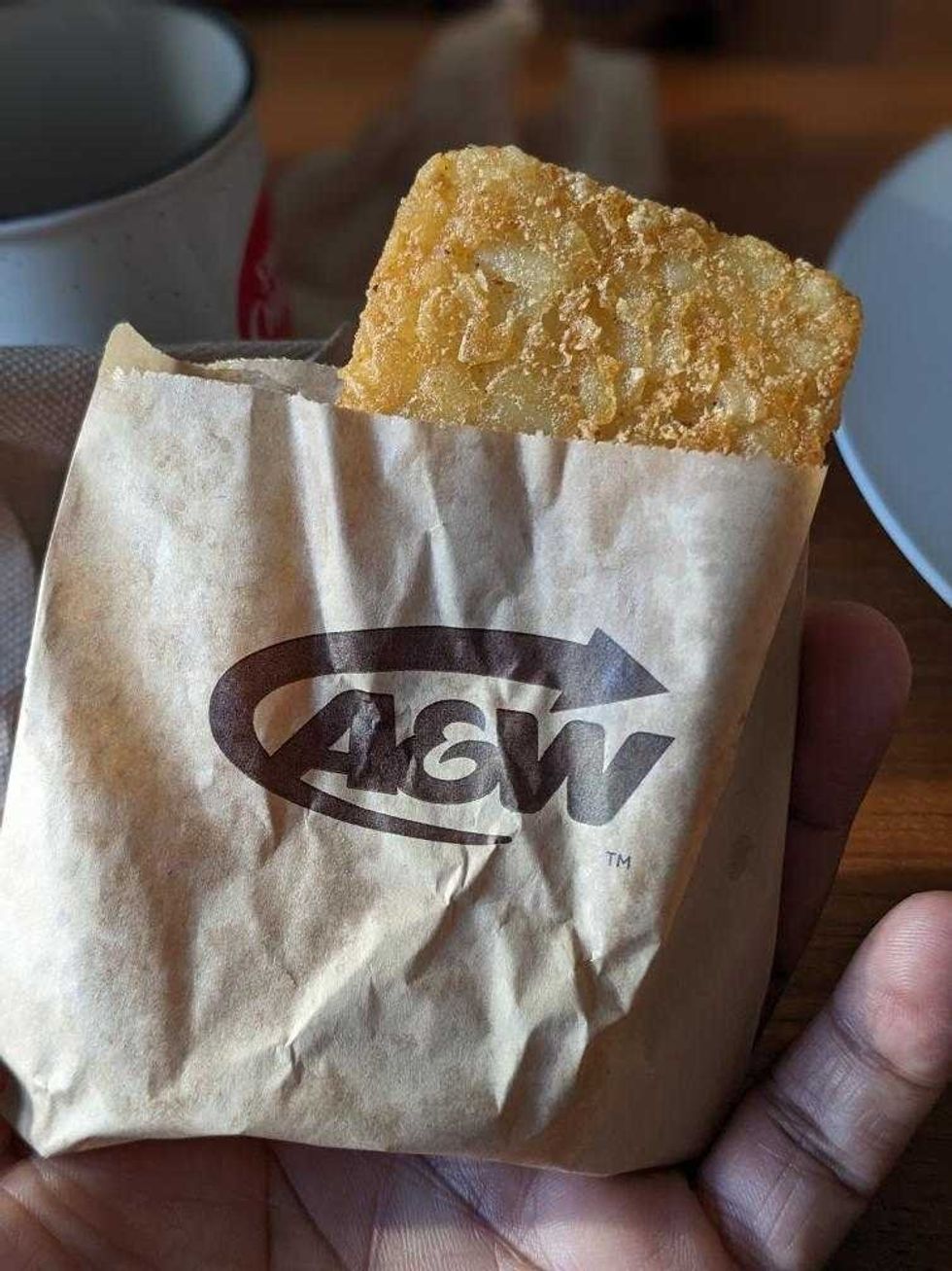A hand holding a brown paper bag with a hash brown peeking out of the top.