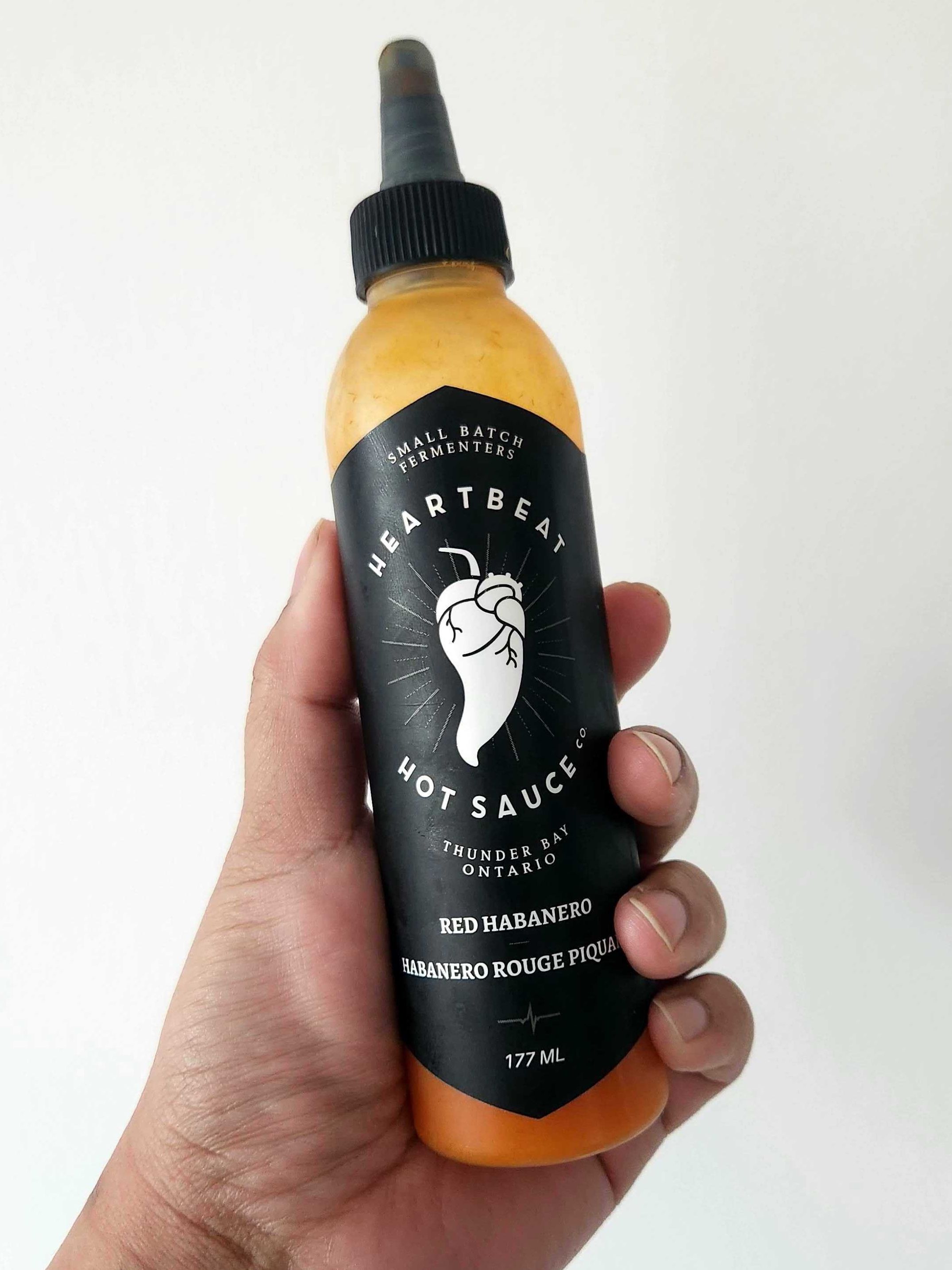 A hand holding a clear bottle of hot sauce with a black label and black lid.