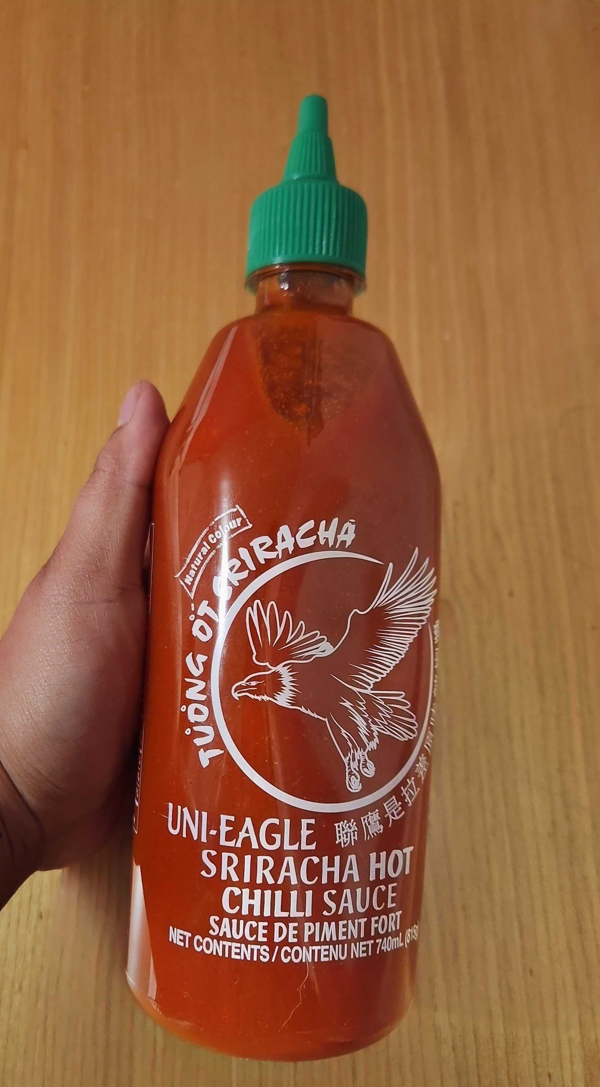 A hand holding a clear bottle of hot sauce with a green cap.