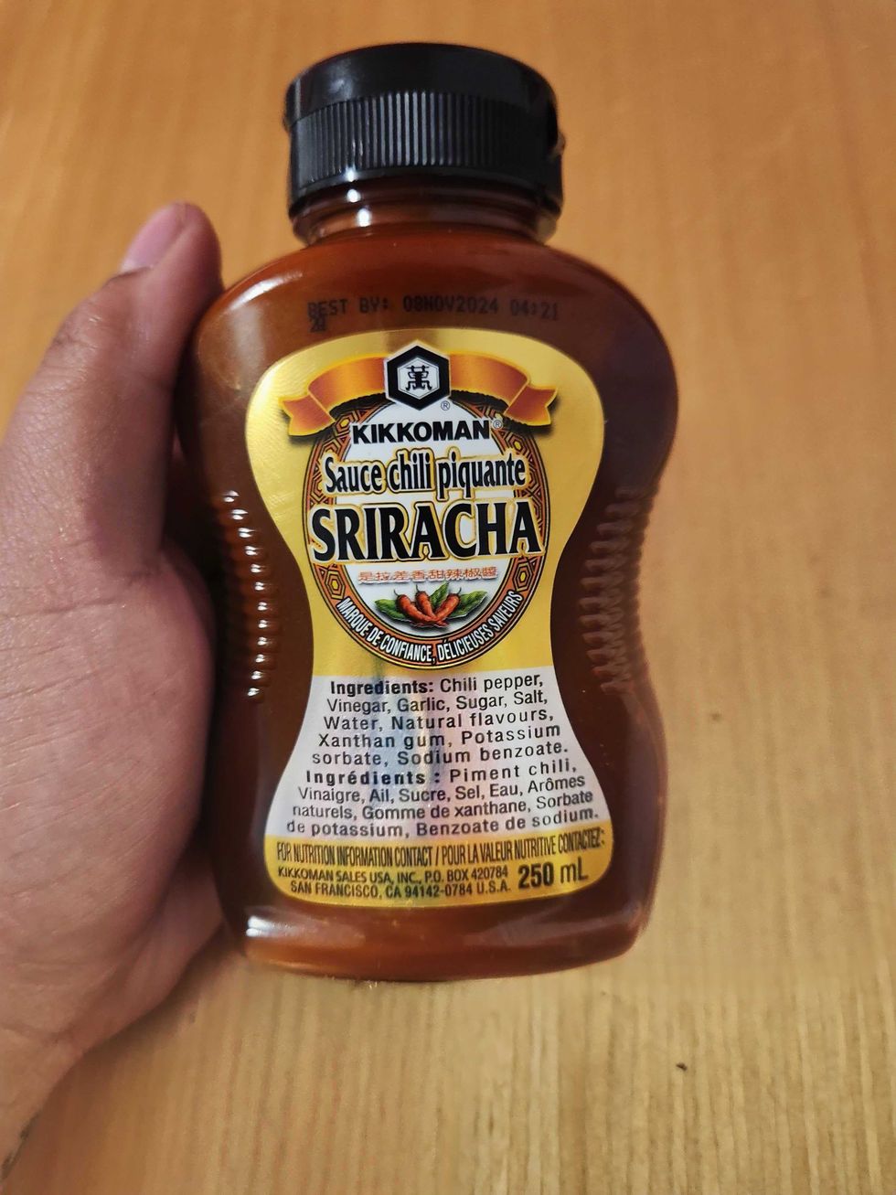 A hand holding a clear bottle of hot sauce with gold label and a black cap.