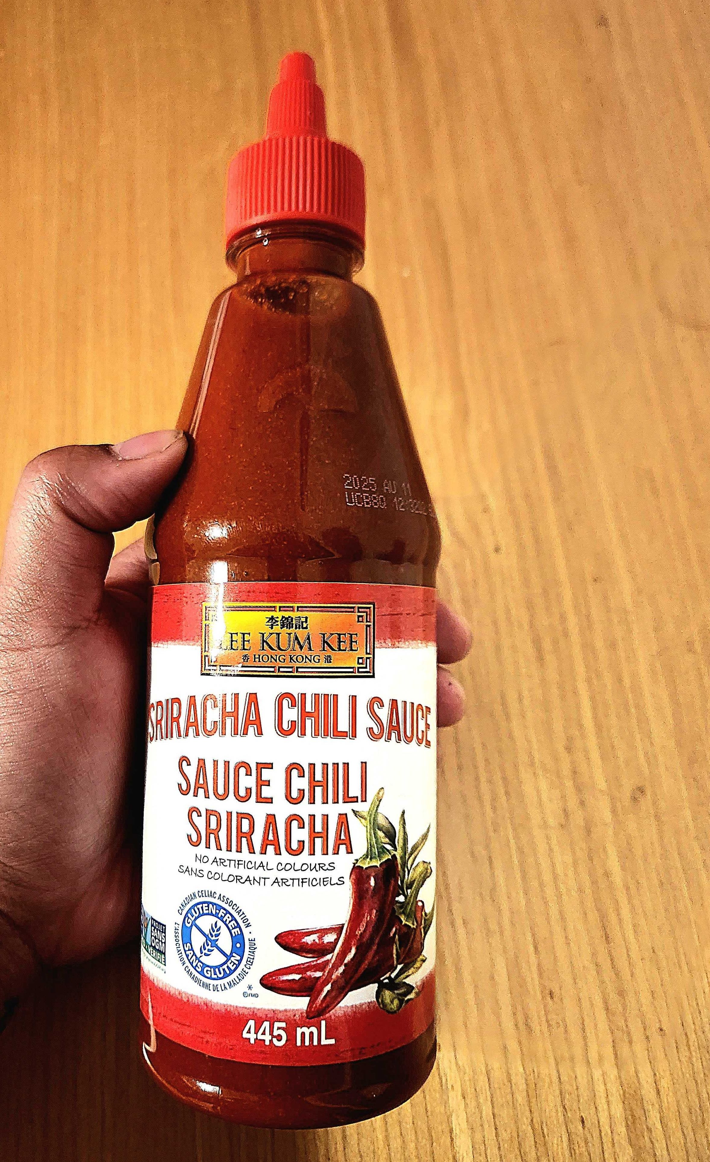 A hand holding a clear bottle of hot sauce with white label and a red cap.