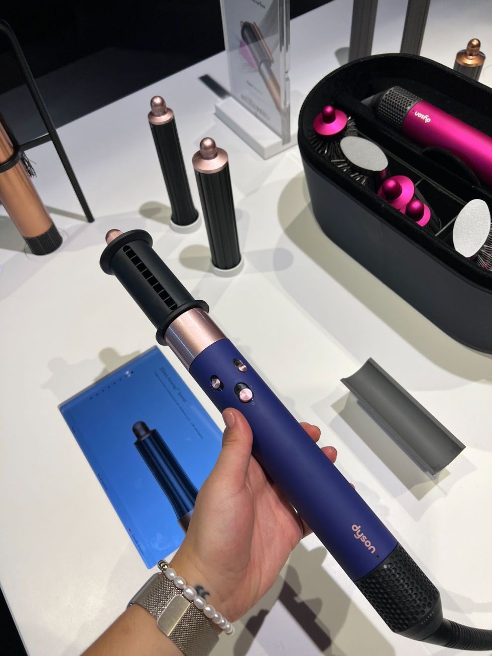 A hand holding a Dyson Airwrap in Prussian Blue and Copper, behind it on the display table are hair tool attachments in black, copper and pink.