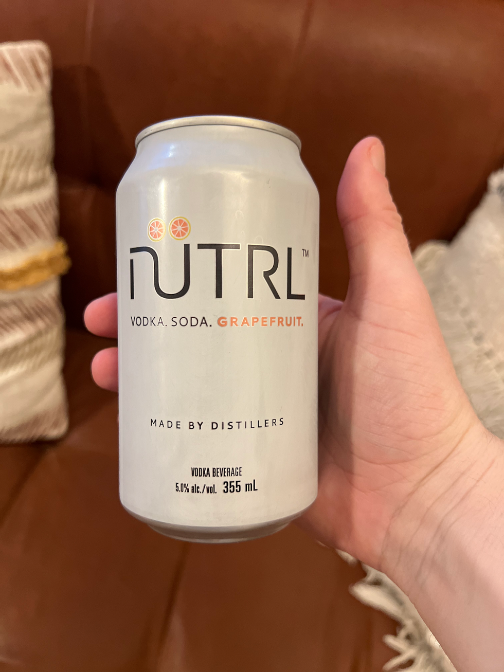 A hand holds a can of N\u00fctrl Grapefruit Vodka Soda in front of a brown leather couch.