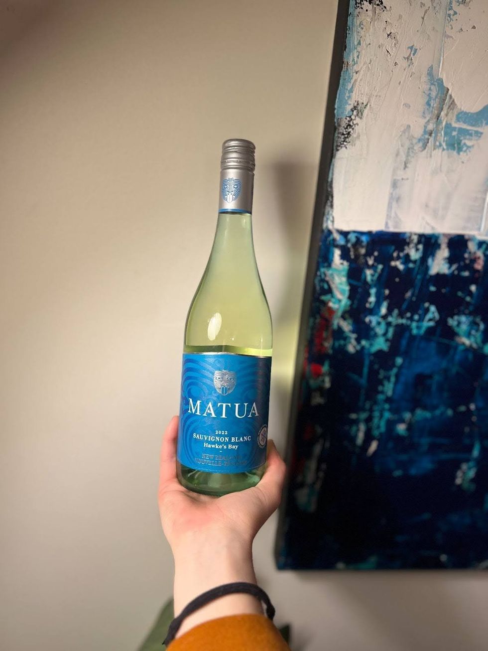 A hand holds out a bottle of Matua Sauvignon Blanc Hawke's Bay in front of a light-coloured wall.