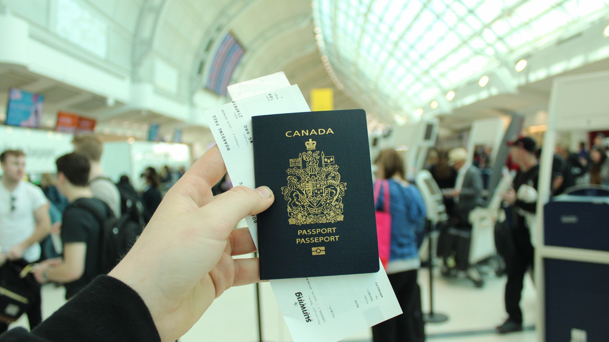 A hand holds up a Canadian passport with an airport in the background.