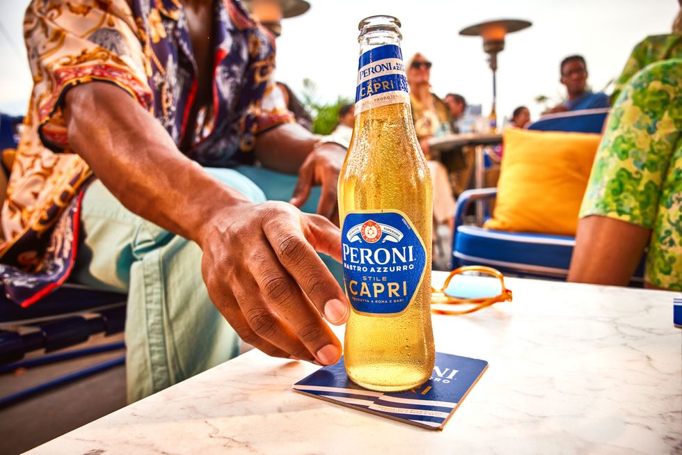 A hand reaches for a bottle of Peroni Nastro Azzurro Stile Capri on an outdoor table.